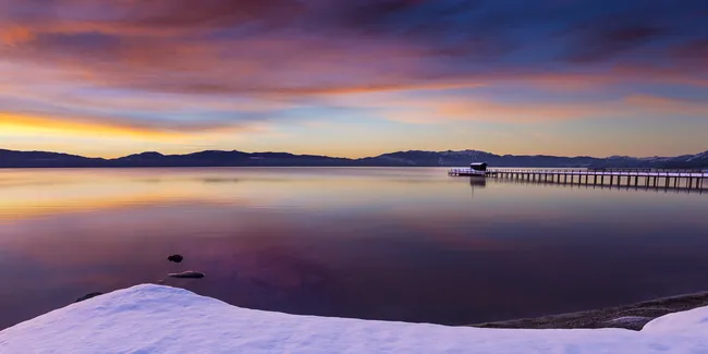 Snow-covered lakeshore at sunrise with calm water, a long wooden pier, and colorful clouds reflecting in the sky.