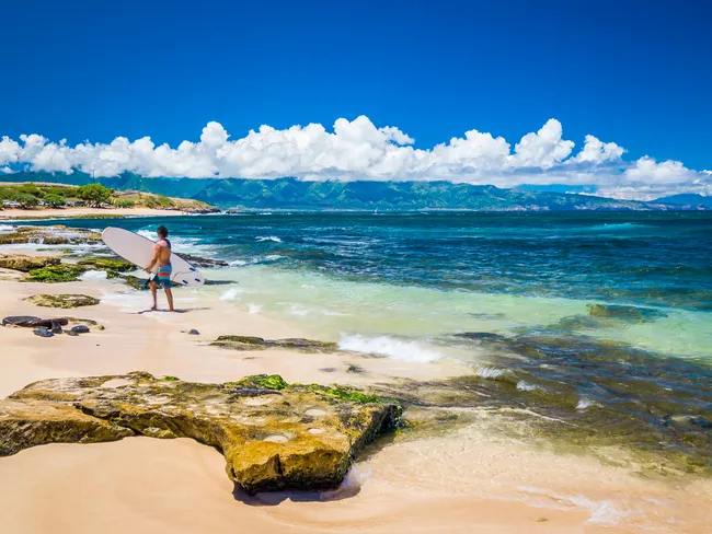 Surfer stands with board on the shore of Hoʻokipa Beach, Maui, with turquoise waters and lush mountains.