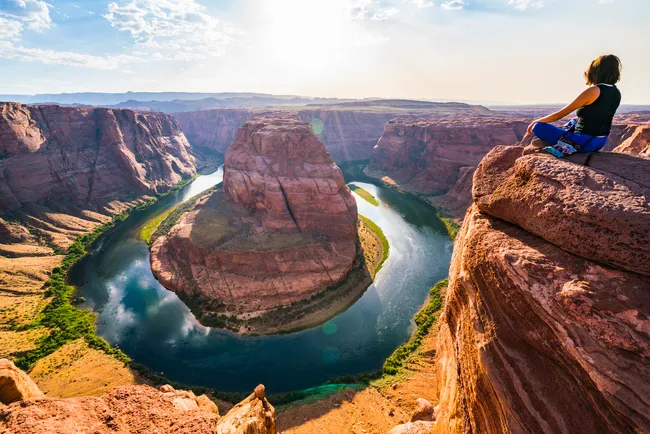 A wide-angle, high-perspective shot of Horseshoe Bend in Arizona. On the right, a person in a black tank top and blue pants sits cross-legged on a large, layered sandstone rock overlooking the deep canyon. Below, the Colorado River makes a smooth, symmetrical 270-degree turn around a massive rock pillar. The canyon walls are bathed in golden afternoon sunlight, and the bright sun glows from the top of the frame, casting a warm haze over the entire desert landscape and the emerald-blue water below.