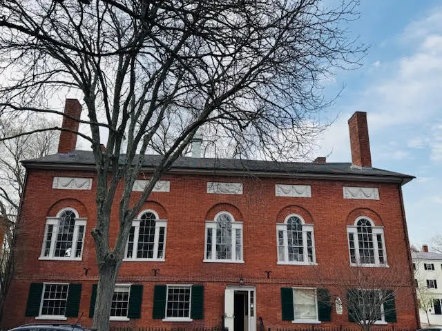 Red-brick Federal-style building with tall arched windows and green shutters, partially obscured by leafless tree in front.