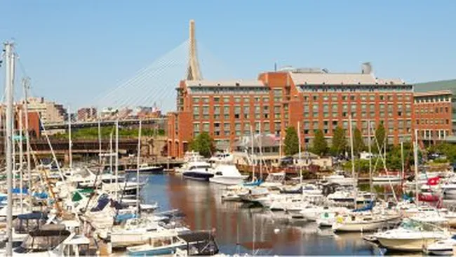 Marina filled with docked sailboats and yachts beside red brick buildings and a cable-stayed bridge under a clear sky.
