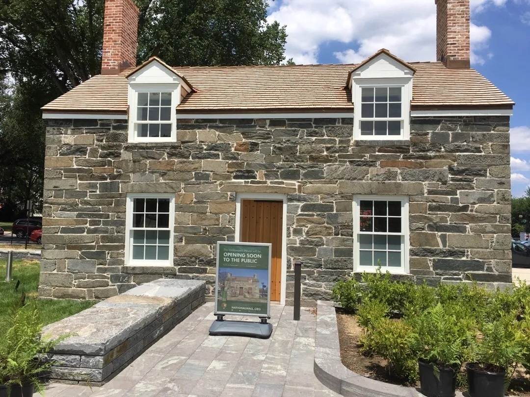 Exterior of the Lockkeeper's House, a small historic two-story stone cottage on the National Mall in Washington, D.C. The building features rough-cut stone walls, a cedar-shingled roof with two dormer windows, two brick chimneys, white-trimmed windows, and a wooden front door. A sandwich board sign in front reads "The Lockkeeper's House will be Opening Soon to the Public — NationalMall.org." The entrance is framed by a newly landscaped stone pathway with freshly planted shrubs. Trees and a partly cloudy sky are visible in the background.