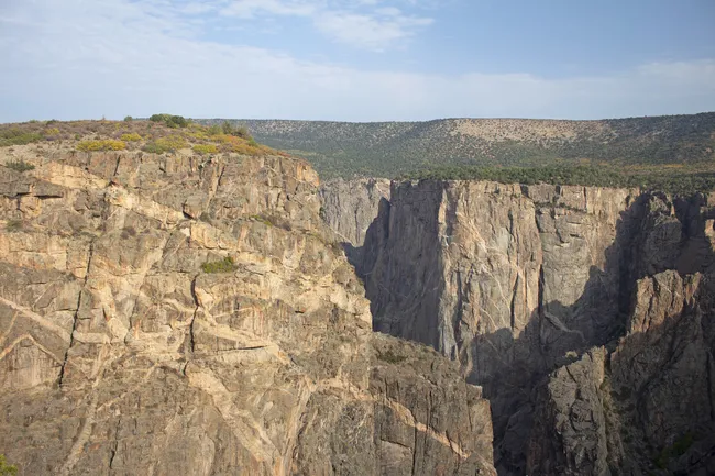 Massive rock cliffs at Black Canyon of the Gunnison rise sharply under a soft morning sky, revealing rugged textures and dramatic vertical drops.