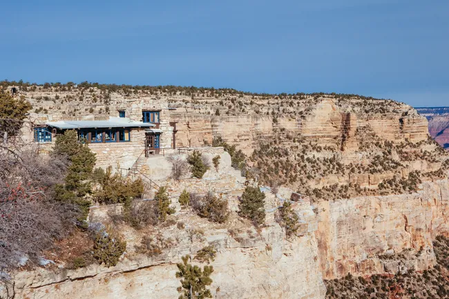 The historic Yavapai Geology Museum, a stone building perched precariously on the rugged edge of the Grand Canyon's South Rim under a bright blue sky.