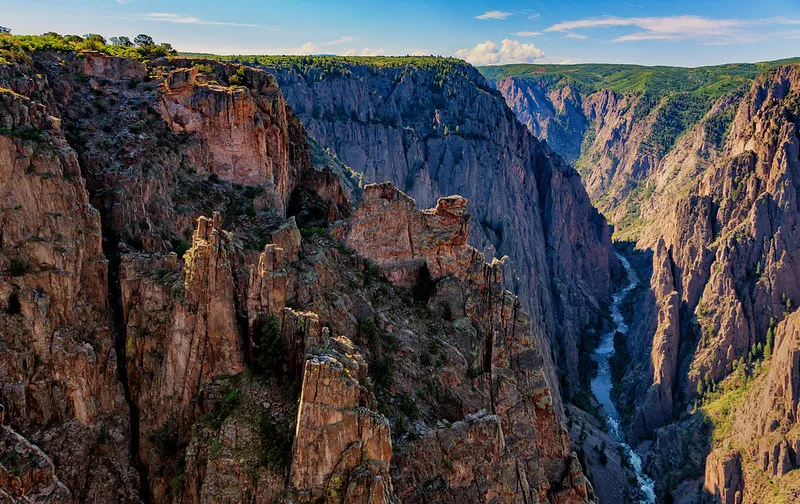 Jagged red rock cliffs drop sharply into the narrow, shadowed gorge of Black Canyon, with a winding river far below and sunlit green hills in the distance.