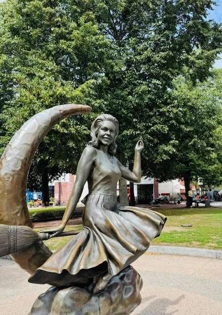 Statue of Samantha from the TV show Bewitched, posed mid-twirl on a crescent moon, set in a park with lush green trees in the background.