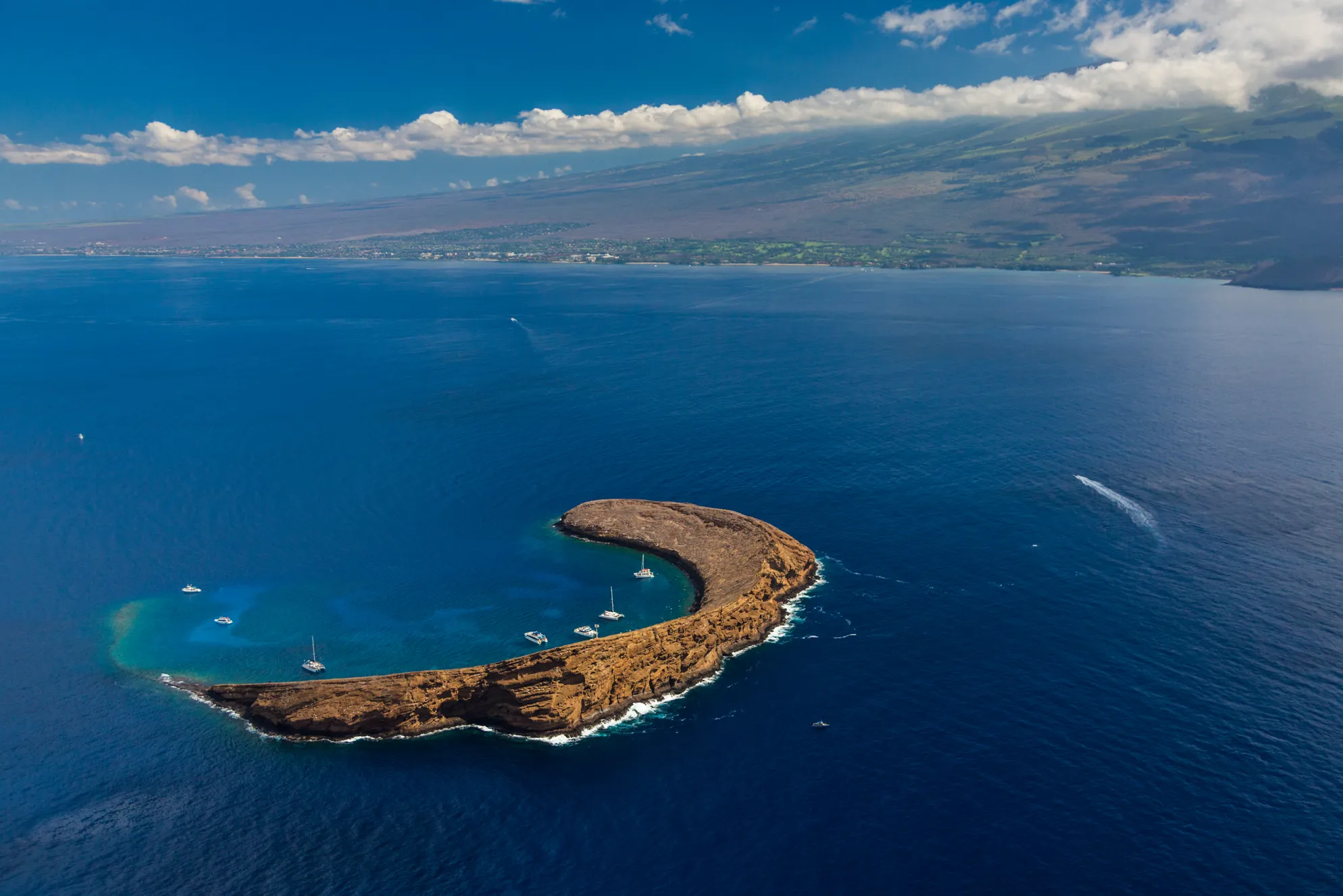 Aerial view of Molokini Crater surrounded by deep blue ocean with several boats anchored inside the crescent-shaped reef.