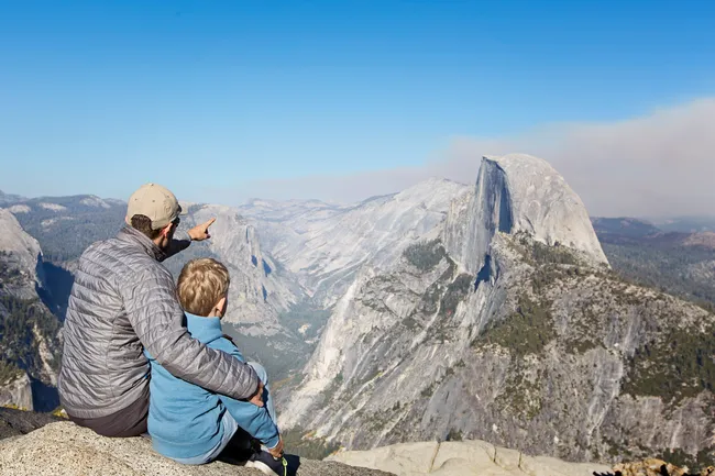 A person and a child overlook Yosemite Valley, with Half Dome visible in the background on a clear day.