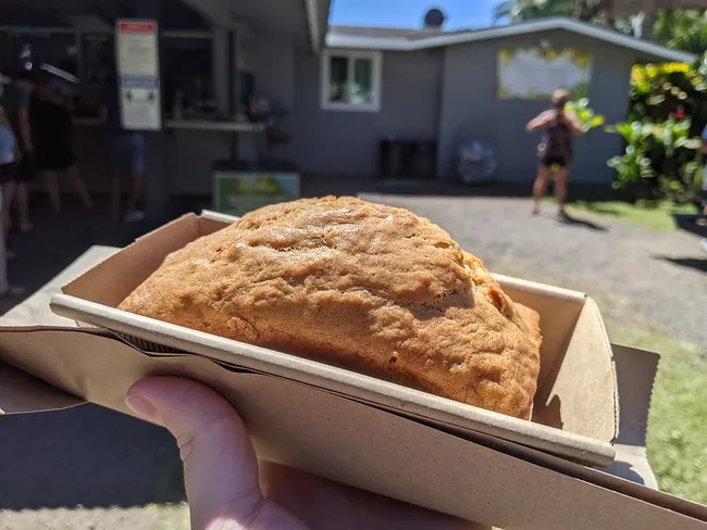 A fresh, golden-brown loaf of banana bread held in a cardboard tray outside a food stand on a sunny day.