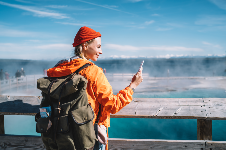 A woman wearing a bright orange jacket and a red beanie stands on a wooden boardwalk, holding a smartphone. In the background, white steam rises from a bright turquoise geothermal pool under a clear blue sky.