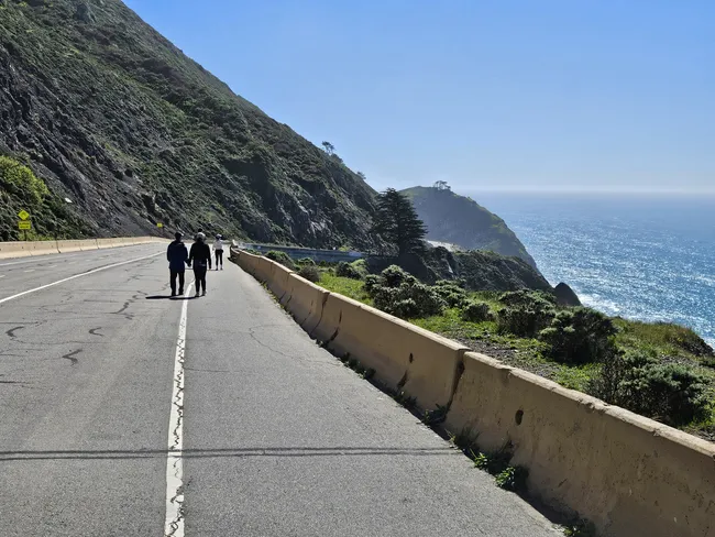 A paved coastal roadway winds along steep green cliffs overlooking the sparkling ocean under a cloudless blue sky. Several people walk along the road beside a concrete barrier, with dramatic seaside bluffs and rugged shoreline stretching into the distance.