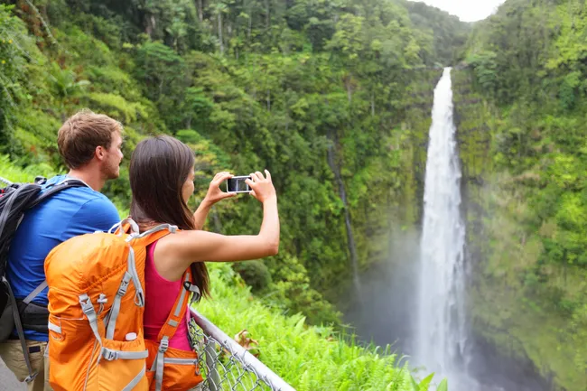 Two hikers with backpacks take photos of Akaka Falls, a tall waterfall surrounded by lush rainforest.