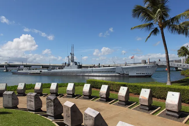 Historic submarine docked at Pearl Harbor, viewed from a memorial site with engraved stone markers and palm trees.
