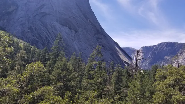 A towering granite cliff rises sharply above a dense forest of pine trees, with a distant waterfall spilling down a rocky canyon under a bright, hazy sky.