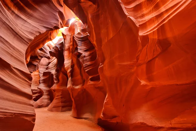 A mesmerizing interior shot of a slot canyon with smooth, undulating sandstone walls. The rock is a vibrant palette of deep orange, red, and burnt sienna, carved into fluid, wave-like shapes by water erosion. Soft, indirect sunlight filters down from the narrow opening above, creating a warm glow that illuminates the sandy floor and highlights the layered textures of the canyon walls.
