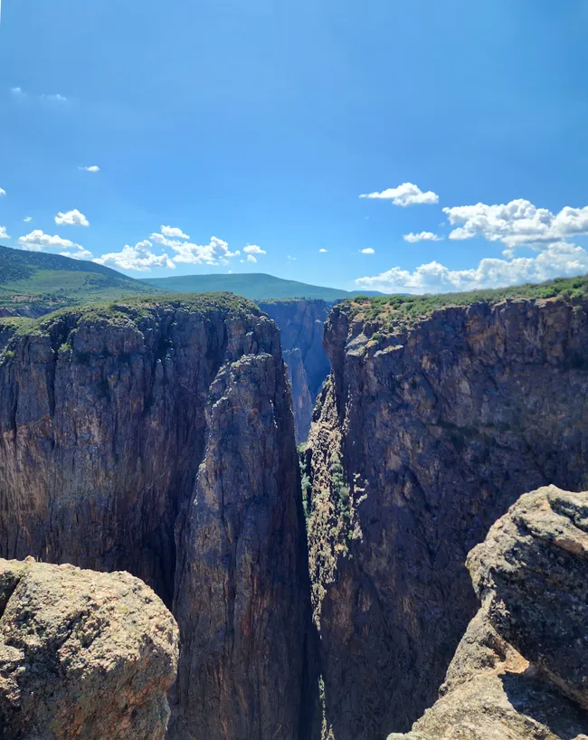 View from the canyon rim at Black Canyon of the Gunnison showing steep vertical cliffs and a narrow chasm under a bright, sunny sky with scattered clouds.