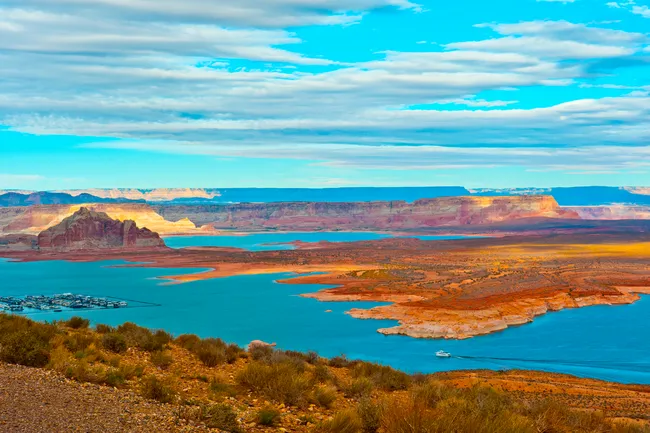 A wide-angle, high-perspective landscape of Lake Powell. The bright turquoise water of the reservoir winds between jagged, flat-topped red sandstone mesas and desert plateaus. In the distance, a small cluster of boats at a marina is visible, and a lone white boat leaves a wake across the water. The sky is a vibrant blue with horizontal layers of wispy white clouds. The desert foreground is dotted with brown and green sagebrush.