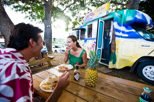 Couple enjoying Hawaiian plate lunch at a colorful food truck under the shade of large trees.
