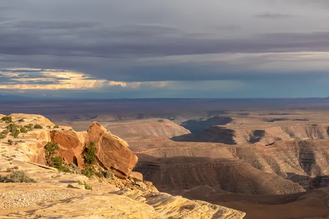 Vast desert canyon view from Muley Point with dramatic cliffs, golden rock ledges, and moody evening skies.