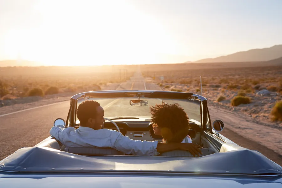 A couple cruises through a desert highway in a convertible at golden hour, surrounded by arid scenery