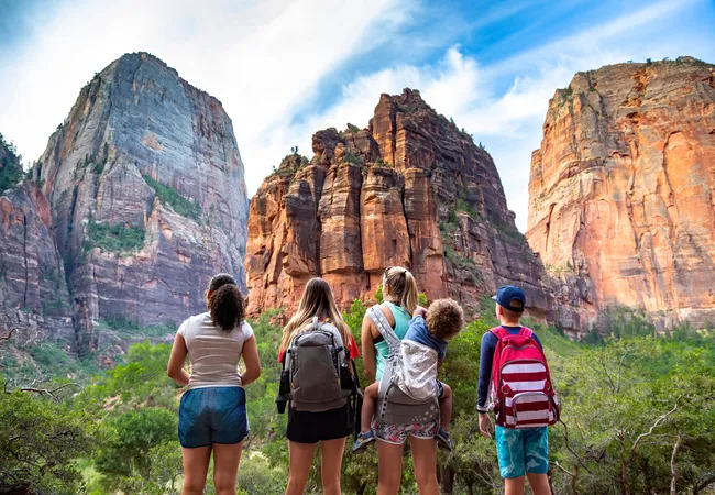 A group of five people with backpacks stands on a trail, facing towering red-and-orange canyon walls that rise above a green valley.