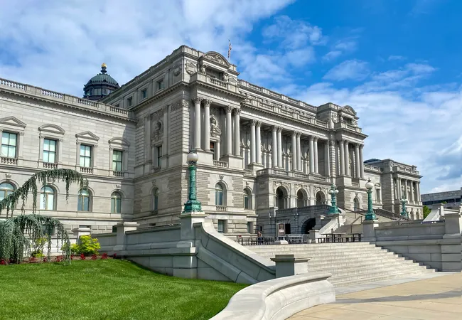 A side-angle exterior view of the Library of Congress Thomas Jefferson Building in Washington, D.C., on a sunny day. The grand Beaux-Arts granite facade features ornate sculptural details, arched windows, Corinthian columns, and decorative green bronze lamp posts flanking the wide stone staircase leading to the entrance. A weeping willow tree and red flower beds are visible on the left, while a lush green lawn fills the foreground under a partly cloudy blue sky.