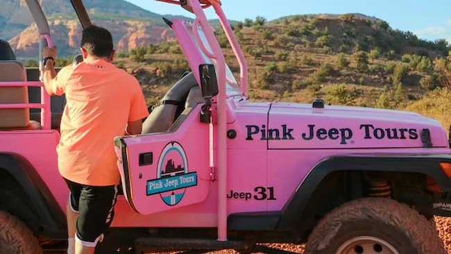 A man wearing an orange shirt steps into a bright pink off-road Jeep with “Pink Jeep Tours” branding.