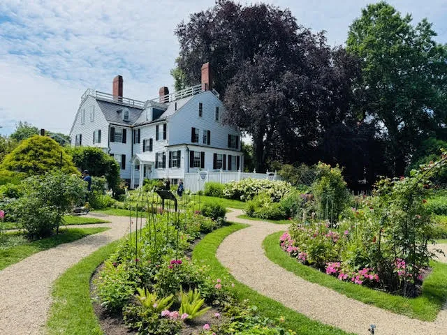 Back view of the historic Ropes Mansion with vibrant flower gardens and winding gravel paths in the foreground.