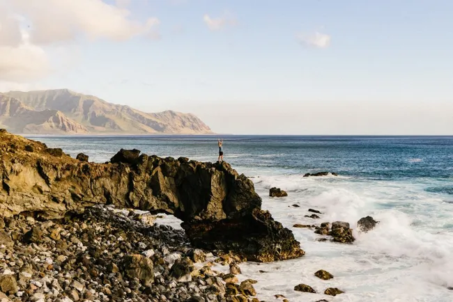 Person stands triumphantly on a rocky cliff overlooking the ocean with mountains in the background. Waves crash below, creating a dramatic coastal scene.