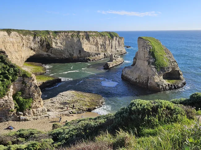A dramatic coastal cove features towering sandstone cliffs, sea caves, and a striking rock formation rising from the water just offshore. Clear blue ocean waters surround the rugged shoreline, while visitors explore the small beach and rocky tide-worn platforms below.