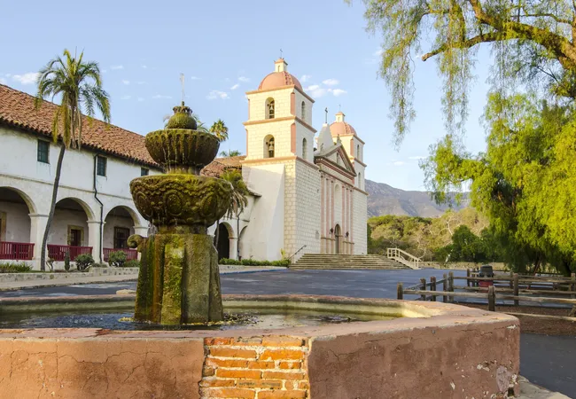 A wide, daytime shot of the historic Old Mission Santa Barbara. In the foreground is a large, weathered stone fountain covered in green moss. The mission building in the background features two white bell towers with pink-domed tops, a grand stone staircase, and a series of arched walkways under a red-tiled roof. A tall palm tree and lush green foliage frame the scene under a clear blue sky, with soft mountains visible in the distance.