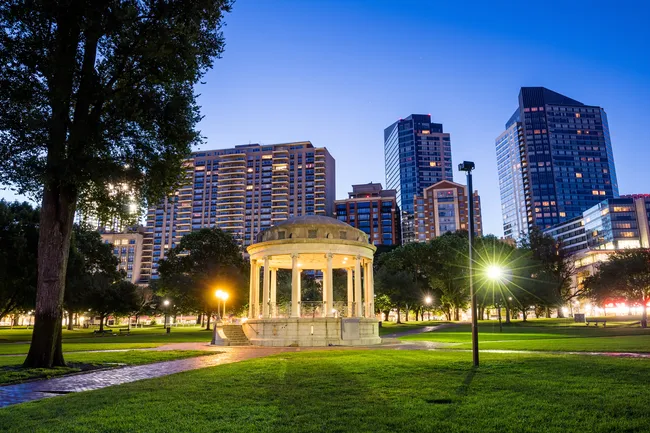 City park at dusk with a lit stone gazebo surrounded by green grass, trees, and tall modern buildings.
