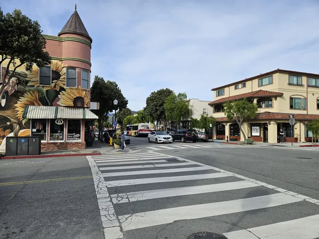 A small-town downtown intersection features colorful storefronts, parked cars, and pedestrians crossing a marked crosswalk. A corner building with a round tower is decorated with a large sunflower mural, adding character to the streetscape beneath an overcast sky.
