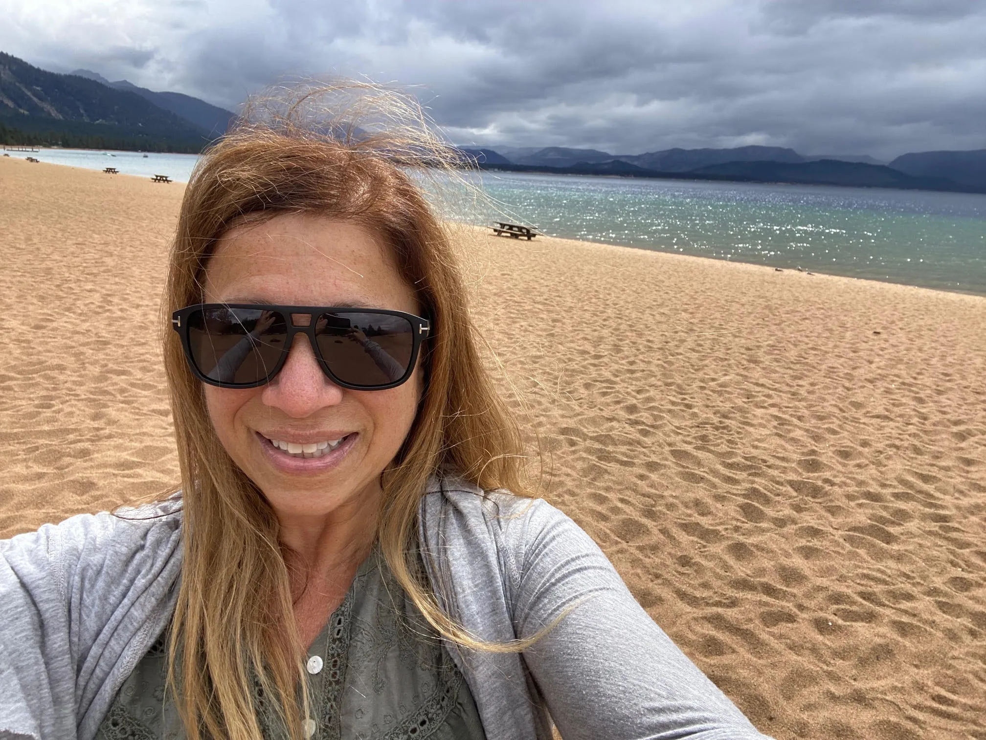 Woman taking a selfie on a wide sandy beach with mountains and Lake Tahoe in the background under cloudy skies.