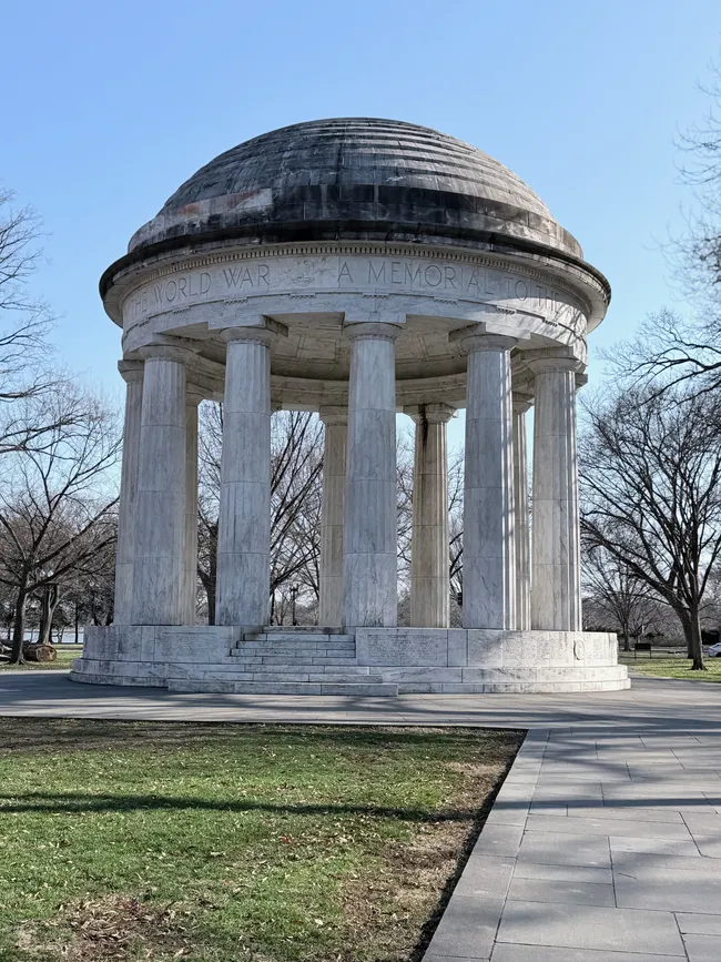 The District of Columbia World War I Memorial, a circular neoclassical white marble domed temple structure on the National Mall in Washington, D.C. The open-air rotunda features tall fluted columns surrounding a central domed roof with a darkened stone surface. An inscription along the frieze reads "The World War — A Memorial to the..." Names of fallen servicemembers are engraved along the base. Steps lead up to the open interior. Bare winter trees frame the structure on both sides under a clear blue sky.
