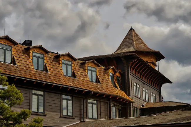 A medium shot of the upper exterior of the historic El Tovar Hotel under a moody, overcast sky. The building features dark brown wood siding and a steep, cedar-shingled roof with several gabled dormer windows. On the right, a distinctive wood turret with a pyramidal roof rises above the structure, supported by ornate dark-red wooden brackets. Large, dark-framed windows are visible across the facade, and the top of a green pine tree is seen in the lower left corner.