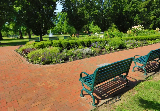 A sunny summer scene in Schiller Park in German Village, Columbus, Ohio. Two green metal park benches sit along a curved red brick pathway beside lush garden beds filled with colorful flowers, lavender, and trimmed boxwood shrubs. Additional benches and a small water fountain spraying in the background are visible amid a canopy of tall, leafy green trees.