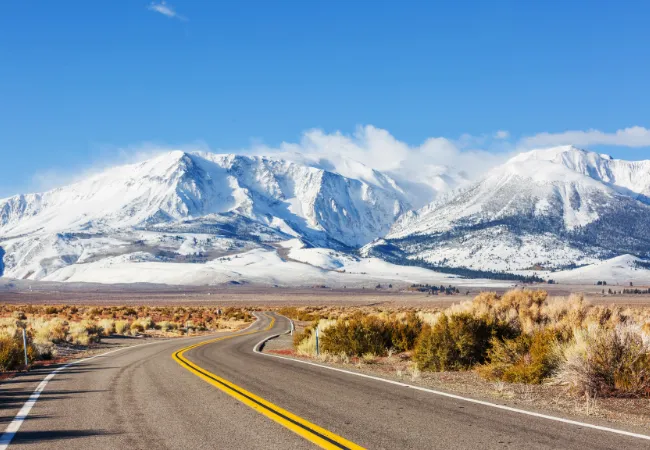 A winding highway cuts through desert scrub with the snow-covered Sierra Nevada mountains rising majestically in the distance.