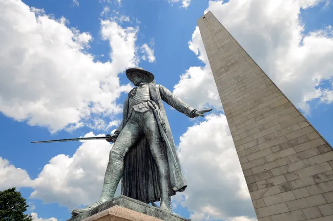 A bronze statue of a Revolutionary War soldier stands before a tall stone obelisk under a partly cloudy sky.