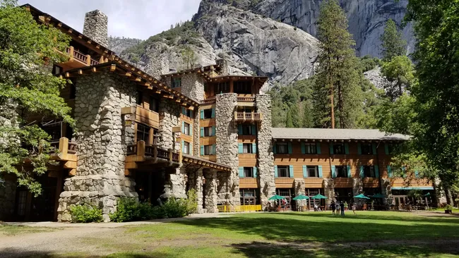 A grand, multi-story lodge built from dark wood and massive grey stones features several balconies and a large stone chimney. The building sits on a manicured green lawn at the base of a towering, sheer granite mountain face, with tall evergreen trees and green patio umbrellas visible near the entrance.