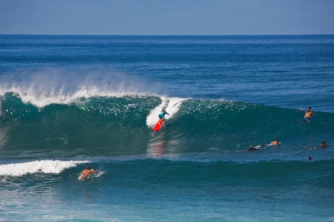 Surfer riding a powerful wave on a red board at a crowded beach break with deep blue ocean backdrop.