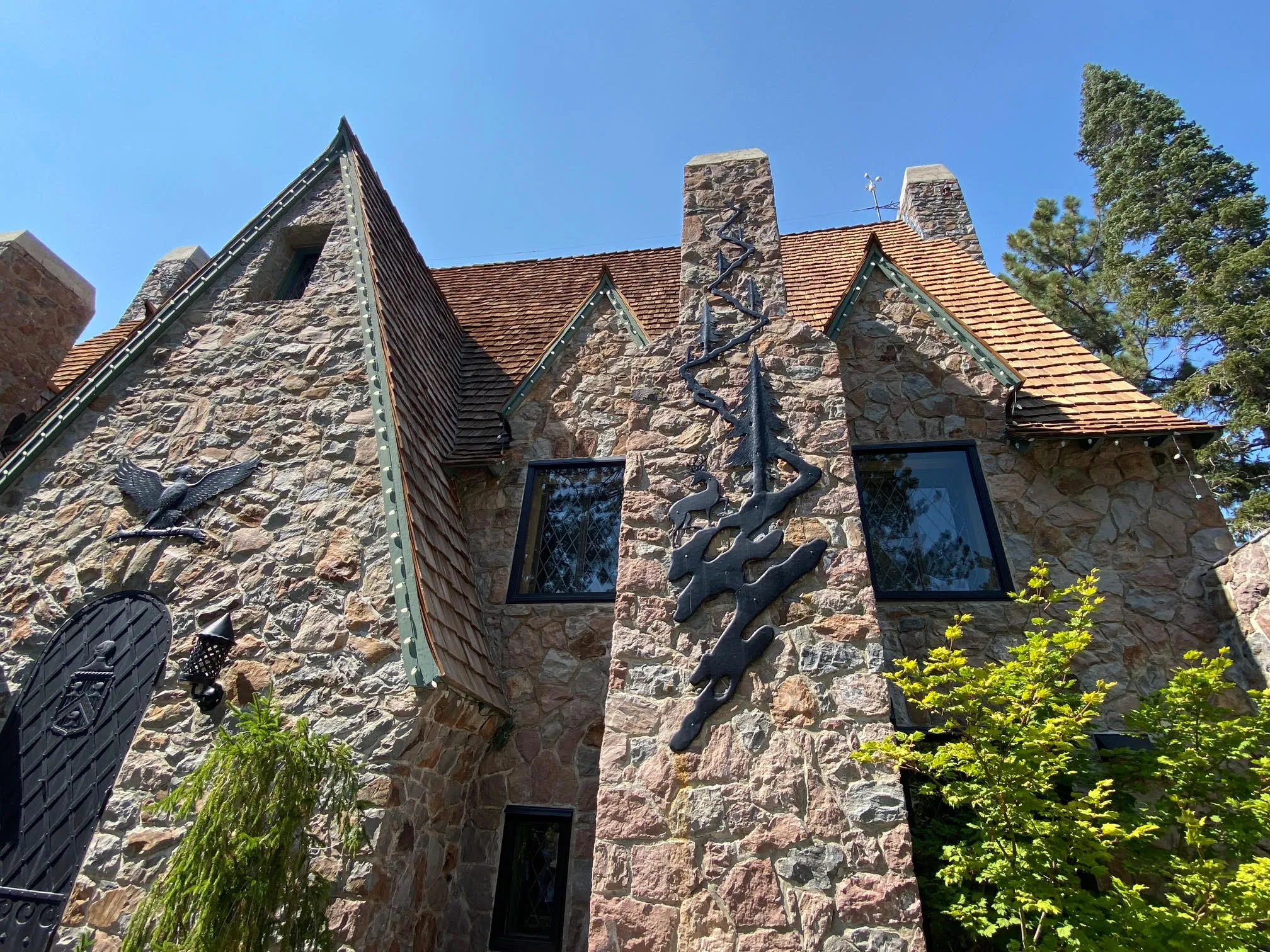 A rustic stone mansion at Thunderbird Lodge features steep gabled roofs, iron artwork on the chimney, and surrounding trees under a clear blue sky.
