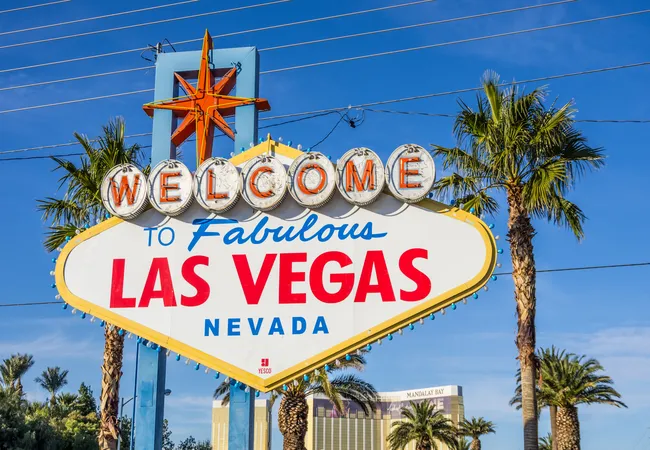 A retro-style roadside sign with bright lights and bold lettering stands among palm trees under a clear blue sky.