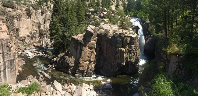 A waterfall cascades between towering granite cliffs into a rocky riverbed, surrounded by dense pine forest in a rugged canyon setting at Shell Falls, Wyoming.