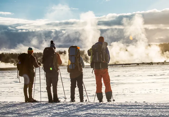Four winter hikers stand in the snow with backpacks and poles, watching steam rise from distant geysers in the cold morning light.