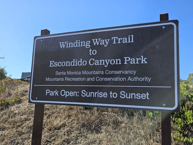 A close-up shot of a dark brown park sign with white text that reads "Winding Way Trail to Escondido Canyon Park." The sign also lists the Santa Monica Mountains Conservancy and the Mountains Recreation and Conservation Authority, with "Park Open: Sunrise to Sunset" at the bottom. The sign is set against a background of dry, golden-brown hillside grass under a clear blue sky.