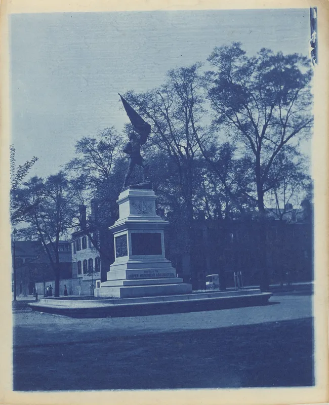 Monument in a public square surrounded by trees, with historic buildings in the background.