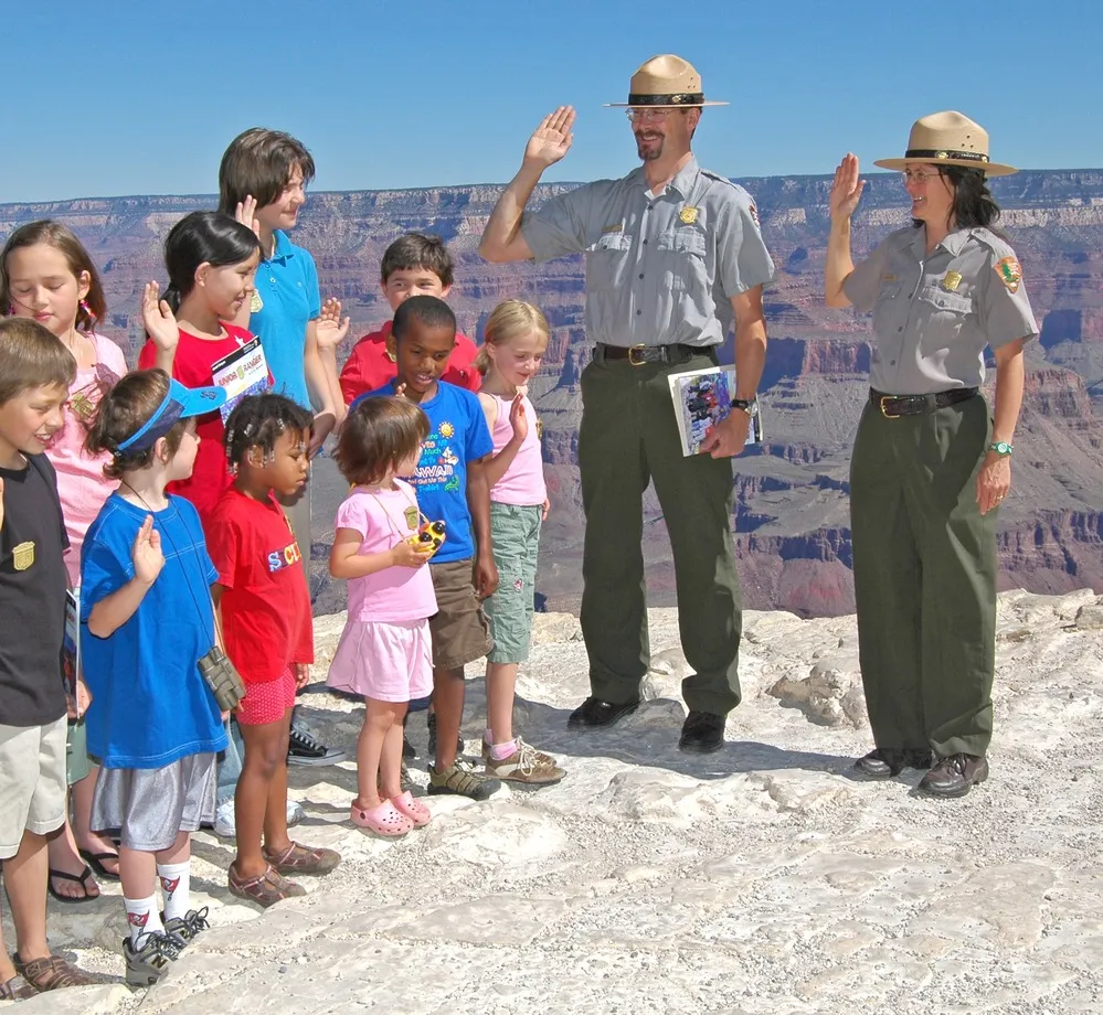 Two National Park Rangers leading a diverse group of children in a Junior Ranger swearing-in ceremony on the rocky edge of the Grand Canyon South Rim.