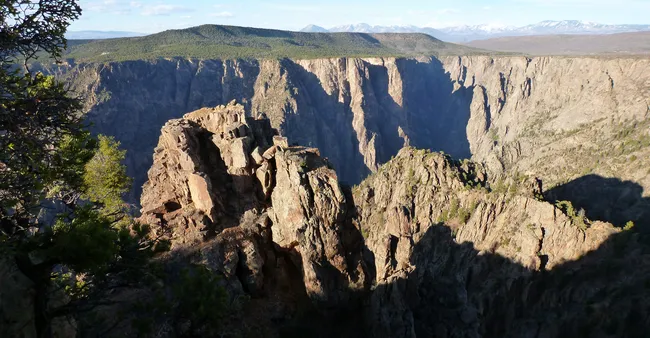 Jagged rock formations in Black Canyon cast deep shadows, with steep cliffs and forested mesas beyond.