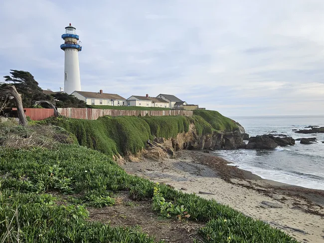 A tall white lighthouse rises above a rugged stretch of coastline, standing beside historic buildings on a bluff covered in lush green vegetation. Below, waves wash onto a narrow rocky beach, while the ocean extends to the horizon beneath a soft, overcast sky.